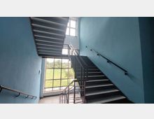 Treppenhaus mit blauen Wänden, schwarzen Handläufen und einer großen Fensterfront, die Tageslicht hereinlässt und den Blick auf eine grüne Wiese ermöglicht.