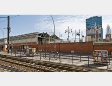 Straßenbahnhaltestelle mit Bahngleisen im Vordergrund, vor einer roten Backsteinmauer und einem historischen Bahnhof mit Metall- und Glasdach. Im Hintergrund sind eine moderne Bürogebäude-Fassade und Straßenlaternen zu sehen.