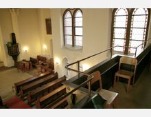 Innenansicht einer Kirche mit Holzbänken und Stühlen, flankiert von schmuckvollen, bunt verglasten Fenstern. Eine hölzerne Kanzel und dezente Wandleuchten ergänzen das schlichte, helle Interieur.