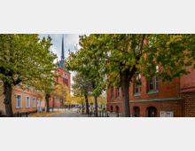 Straßenszene mit Backsteinarchitektur, Bäumen im Herbstlaub und einer Kirche mit rotem Turm und hohem, spitzen Dach im Hintergrund. Links und rechts der gepflasterten Straße stehen Gebäude aus rotem und gelbem Mauerwerk.