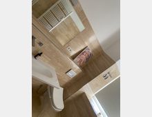 Modernes Badezimmer mit beigefarbenen Fliesen an Wänden und Boden, einem weißen Wand-WC und einem rechteckigen Waschbecken. Im Hintergrund befindet sich ein großer Spiegel, eine elektrische Handtuchheizung und ein Fenster mit Rollladen; ein gerahmtes Bild steht auf der Fensterbank.