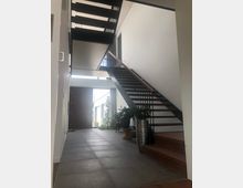 Moderner Innenraum mit Flur und schwebender Treppe aus dunklem Metall mit Holzhandlauf, die nach oben führt. Der Boden besteht aus großen grauen Fliesen, und Pflanzen in Töpfen schmücken den Raum; durch eine geöffnete Tür ist ein Blick auf einen Außenbereich mit Tageslicht möglich.