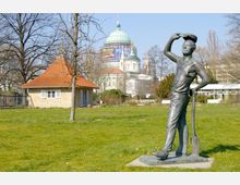 Eine Grünfläche mit einer Bronzestatue eines Arbeiters, der eine Schaufel hält, im Vordergrund. Im Hintergrund sind ein kleines Backsteingebäude mit rotem Ziegeldach und die grünliche Kuppel eines großen, historischen Gebäudes sichtbar.