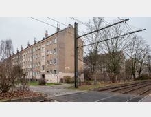 Mehrstöckiges, schlichtes Wohngebäude mit Putzfassade in einem Wohngebiet. Im Vordergrund befinden sich Straßenbahnschienen, kahle Bäume und ein Gehweg.