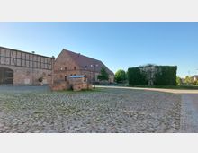 Historischer Hof mit Kopfsteinpflaster, einem großen Backsteingebäude mit Satteldach im Hintergrund und einem Fachwerkgebäude an der linken Seite. Im Mittelpunkt steht ein kleiner, verfallener gemauerter Brunnen, daneben eine begrünte Wandstruktur.