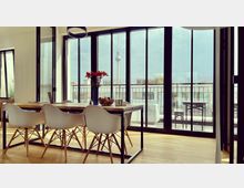 Moderner Essbereich in einer Wohnung mit Holzboden, einem großen Holztisch, weißen Stühlen und einer Vase mit roten Blumen. Im Hintergrund sind bodentiefe Fenster mit Blick auf eine Terrasse und den Berliner Fernsehturm sichtbar.