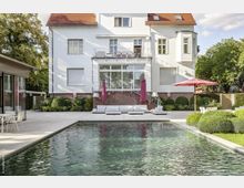 Hinterhof eines modernen Herrenhauses mit weißer Fassade, roten Dachziegeln und großen Fenstern, ergänzt durch eine Terrassenanlage mit Lounge-Möbeln, einem Pool und gepflegter Bepflanzung. Im Vordergrund steht ein Sonnenschirm, und die Umgebung ist von Bäumen gesäumt.