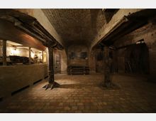 Ein Kellerraum mit Gewölbedecke aus Ziegelsteinen, rustikalen Pfeilern und einer gepflasterten Bodenfläche. Im Hintergrund befinden sich gestapelte Holzpaletten, alte Werkzeuge und vereinzelt beleuchtete Nischen mit Werkgegenständen.