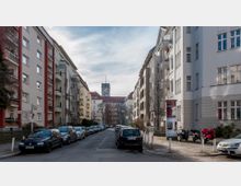 Wohnstraße in einer Stadt mit parkenden Autos links und rechts der Straße. Umgeben von mehrstöckigen Wohngebäuden, führt die Straße zu einem größeren Gebäude mit Turmuhr im Hintergrund.