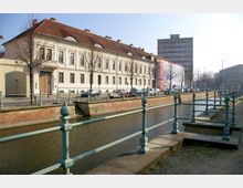 Historisches Gebäude mit heller Fassade und rotem Ziegeldach, gelegen an einem Kanal mit schmiedeeisernem Geländer im Vordergrund; im Hintergrund sind moderne Bürogebäude sichtbar.