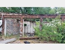 Ruine eines Backsteingebäudes mit teilweise zerstörten Wänden und ohne Dach, umgeben von Wildpflanzen und Bäumen. Im Vordergrund sind Graffiti, Schutt und überwucherte Vegetation zu sehen.