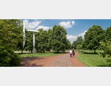 Ein Park mit einem gepflasterten Weg, der von Bäumen und grünen Rasenflächen umgeben ist. Links steht eine moderne Skulptur in Form eines Windrades, und zwei Personen gehen auf dem Weg spazieren.