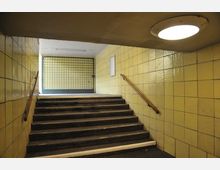 Unterführung mit einer Treppe, flankiert von gelb gefliesten Wänden und Holzhandläufen. Am oberen Ende der Treppe befindet sich ein Gittertor, und eine Deckenleuchte beleuchtet den Bereich.