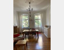 Helles Wohnzimmer mit Holzdielenboden, einem Esstisch aus Holz und vier Stühlen in der Mitte. Die hohen Doppelfenster mit weißen Vorhängen lassen Tageslicht herein, während ein Kronleuchter mit dekorativer Deckenrosette die Decke schmückt. Ein Sideboard steht rechts, links ein bunter Sessel und ein kleiner Kamin.