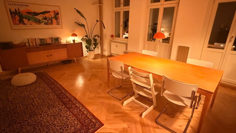 Gemütlicher Essbereich in einer Wohnung mit großem Holztisch, weißen Stühlen und orangefarbenen Pendelleuchten über dem Tisch. Helle Wände, Parkettboden im Fischgrätmuster, großer Teppich, Sideboard mit Büchern, Zimmerpflanze sowie mehrere hohe Fenster und eine Balkontür prägen den Raum.
