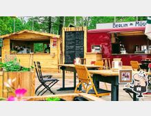 Ein Außenbereich eines kleinen gastronomischen Betriebs mit Holzmöbeln und einer Holzhütte, neben einem roten Verkaufswagen. Im Hintergrund befindet sich eine Speisetafel mit einer handgeschriebenen Menüauswahl. Umliegende Pflanzendekoration und grüne Bäume schaffen eine einladende Atmosphäre.