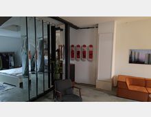 Modern eingerichteter Wohnraum mit Betonboden, Spiegelwänden und einer offenen Schlafzimmerecke links im Hintergrund. Rechts befindet sich ein orangefarbenes Sofa, daneben eine Pflanze, ein Sessel und Wanddekoration mit roten Designelementen sowie ein Wandbild mit Hochhäusern.