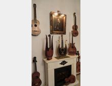 Eine Wand mit verschiedenen Saiteninstrumenten, darunter Gitarren und Lyren, die um einen dekorativen Kamin arrangiert sind. Darüber hängt ein vergoldetes Porträt eines Mannes mit einem Musikinstrument.