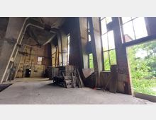 Industriehalle mit hohen, teilweise verfallenen Wänden aus Beton und Ziegeln, großen Fenstern und gesammelten Bauteilen wie Holzplanken und Metallteilen. Durch die geöffneten Fenster ist draußen dichtes Grün zu sehen.