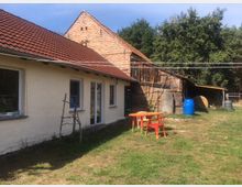 Ein ländliches Grundstück mit einem niedrigen Gebäude mit weiß verputzten Wänden und rotem Ziegeldach, daneben eine Scheune aus Holz und Ziegelstein. Im Vordergrund befindet sich eine Grünfläche mit einem orangefarbenen Tisch, roten und orangen Stühlen sowie einer blauen Tonne.