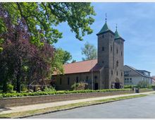Kirche aus Backstein mit zwei Türmen und grün gedeckten Spitzdächern, umgeben von Bäumen und einem niedrig gemauerten Zaun. Im Vordergrund ist ein Gehweg und eine ruhige Straße zu sehen.