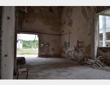 Innenraum eines verlassenen, verfallenen Gebäudes mit abgenutzten Wänden, Graffiti und Schutt am Boden. Durch große Öffnungen in den Wänden ist eine grüne Außenlandschaft und ein weiteres zerfallenes Gebäude sichtbar.