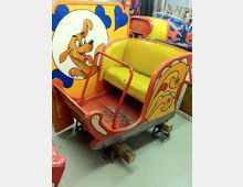 Ein alter Karussellwagen mit gelber Sitzfläche und rotem Rahmen, dekoriert mit bunt gemalten Mustern und dem Bild eines lächelnden Hundes. Der Wagen steht auf Holz- und Metallblöcken in einem Innenraum.