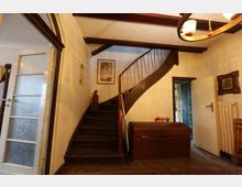 Eingangsbereich eines Hauses mit einer dunkelbraunen, geschwungenen Holztreppe, einem großen Holzkasten und einer Glastür auf der linken Seite. Der Raum ist mit Holzbalken an der Decke, einem Teppichboden und einigen dekorativen Elementen wie einem Bild und einer kleinen Skulptur ausgestattet.