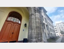 Eingang eines historischen Gebäudes mit einem großen Holztor und einem steinernen Torbogen, flankiert von Gebäudefassaden mit rechteckigen Fenstern; im Hintergrund moderne Wohnhäuser und geparkte Fahrräder entlang eines Gehwegs.