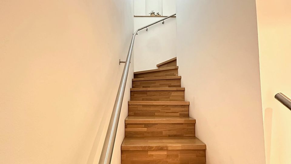 Schmales, modernes Treppenhaus mit Holzstufen und weißen Wänden, entlang einer Seite mit einem Metallhandlauf. Oben sorgt ein hohes, schmales Fenster für Tageslicht im Treppenaufgang.