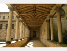 Langer Säulengang mit steinernen Pfeilern und reich verziertem, offenen Holztragwerk, flankiert von sandfarbenen Mauern mit Fenstern und Verzierungen im Hintergrund. Der Boden ist mit hellen Pflastersteinen ausgelegt und der Gang führt zu einer Tür am Ende.