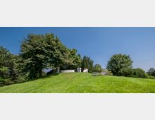 Grüne Parklandschaft mit einem grasbewachsenen Hügel, auf dem mehrere Bäume stehen. Auf dem Hügel befinden sich Steinbänke, eine moderne Liege und eine Person sitzt im Schatten eines großen Baumes.