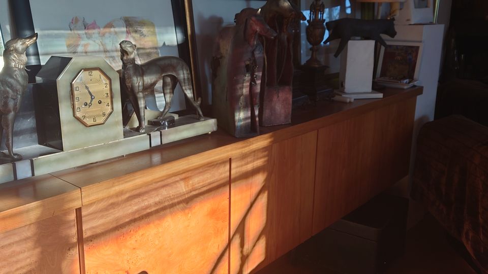 Eine hölzerne Anrichte in einem Raum, dekoriert mit gerahmten Bildern, Metallskulpturen, einer Wanduhr, einem mehrarmigen Kerzenleuchter und einer Tischlampe. Sonnenlicht fällt durch ein Fenster und wirft Schatten auf die Möbel und den Boden.