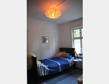 Ein einfach eingerichtetes Schlafzimmer mit einem Einzelbett, das mit einer blauen Decke und gestreiften Kissen dekoriert ist. Ein orange leuchtender Lampenschirm hängt an der Decke, während ein kleines Fenster Tageslicht hereinlässt; daneben steht ein weißer Nachttisch mit einer Lampe.