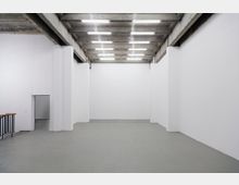 Ein leerer, rechteckiger Raum mit weiß gestrichenen Wänden und einem grauen Boden. Die Decke besteht aus Betonträgern mit eingelassenen LED-Leuchtstoffröhren, und auf der linken Seite befindet sich eine kleine Türöffnung sowie ein Geländer mit hölzernem Handlauf.