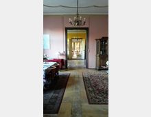 Ein historischer Wohnraum mit rosa Wänden, antiken Möbeln und Kronleuchtern. Im Vordergrund ein Esstisch mit Teppichen auf dem Holzfußboden, im Hintergrund mehrere durchgehende Räume mit weiteren klassischen Möbelstücken.