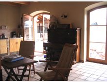 Ein wohnlicher Raum mit Fliesenboden und Holzbalkendecke, ausgestattet mit einem Klapptisch, zwei Stühlen und einem antiken Klavier. Durch große Glastüren fällt Tageslicht herein, und der Blick führt auf eine überdachte Terrasse.