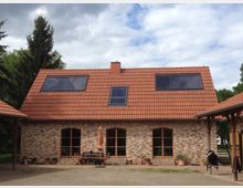 Backsteingebäude mit rotem Satteldach und großen Dachfenstern, umgeben von Bäumen. Vor dem Haus befindet sich eine Terrasse mit Holzmöbeln und einem Sonnenschirm, umgeben von Pflanzkübeln.