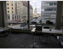 Blick aus einem Büro mit schlicht eingerichteten Schreibtischen durch eine große Fensterfront auf eine Stadtstraße. Im Hintergrund ist der Gendarmenmarkt in Berlin mit dem Deutschen Dom erkennbar, flankiert von historischen und modernen Gebäuden.