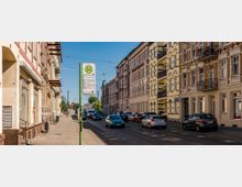 Straßenszene mit mehrstöckigen Altbauhäusern auf beiden Seiten, einem Gehweg, einer Haltestelle mit einem grünen Schild für Busse und Straßenbahnen, parkenden Autos und mehreren Fahrspuren. Im Hintergrund ist blauer Himmel und vereinzelt Vegetation zu sehen.