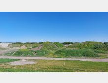 Weite Landschaft mit begrünten Hügeln und vereinzeltem Bewuchs, umgeben von flachen Wegen und Wiesen. Im Hintergrund stehen Windkraftanlagen vor klarem, blauem Himmel.