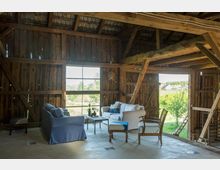 Innenraum einer rustikalen Scheune mit Holzwänden und offenem Dachgebälk. Im Vordergrund steht eine Sitzecke mit Sesseln, Sofas und einem Couchtisch, durch offene Fensteröffnungen ist eine grüne Landschaft und ein Dorf im Hintergrund sichtbar.