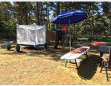 Ein Garten mit vereinzelten Bäumen und Rasenfläche, darauf ein Bett mit Moskitonetz, ein blauer Sonnenschirm, ein Surfbrett auf einem Gestell, eine Schubkarre, ein aufblasbarer Pool und einige Gartenmöbel. Im Hintergrund ist ein Holzzaun zu sehen.