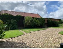 Ein rustikaler Hof mit Kopfsteinpflaster, einer großen Scheune mit rotem Ziegeldach und teilweise mit Efeu bewachsenen Wänden. Umgeben von grüner Wiese, Bäumen und Sträuchern; der Himmel ist bewölkt mit sonnigen Abschnitten.