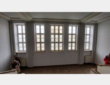 Ein Raum mit hohen weißen Wänden, drei großen Fenstern in der Mitte und zwei schmaleren Fenstern an den Seiten, alle mit dekorativem Glasmuster. Im unteren Bereich sind Personen an einer weißen Treppe mit Holzelementen sichtbar.