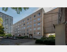 Wohnblock aus Beton in einer städtischen Umgebung mit mehreren Etagen und rechteckigen Fenstern. Im Vordergrund ist eine Straße mit geparkten Autos sowie kleinere Grünflächen zu sehen, im Hintergrund steht ein weiteres, höheres Gebäude.