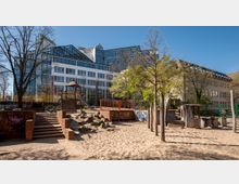 Spielplatz mit Sandfläche, Klettergerüst, Rutsche und Holzhäuschen, umgeben von Bäumen. Im Hintergrund sind moderne Bürogebäude und ältere Gebäude mit großen Fenstern zu sehen.