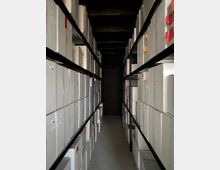 Ein schmaler Lagerraum mit hohen Metallregalen auf beiden Seiten, die mit stapelweise beschrifteten Archivboxen befüllt sind. Der Raum hat eine dunkle Decke und eine einfache graue Bodenfläche.