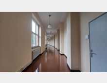 Ein langer, heller Flur mit cremefarbenen Wänden und einer glänzend braunen Bodenfläche. Auf der linken Seite befinden sich große Fenster mit Heizkörpern darunter, und an der Decke hängen regelmäßig angeordnete Hängelampen.