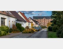 Straßenszene in einem Wohngebiet mit Reihenhäusern auf der linken Seite, die rote Ziegeldächer und gepflegte Vorgärten haben. Im Hintergrund steht ein großes, historisches Backsteingebäude mit geschlossen wirkenden Fenstern, umgeben von Herbstbäumen.